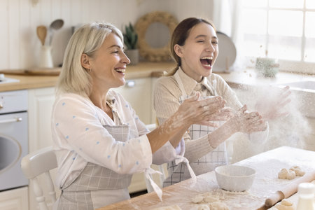 Happy excited teen granddaughter and grandma in aprons having sun, baking in kitchen, throwing flour powder over table with dough, laughing, shouting for joy, enjoying cooking entertainmentの写真素材