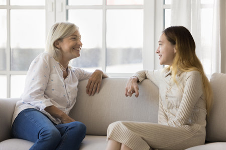 Positive teenage child and happy grandmother sitting on couch together, relaxing at home, talking, laughing, having fun. Grandma and teen granddaughter meeting at homeの写真素材