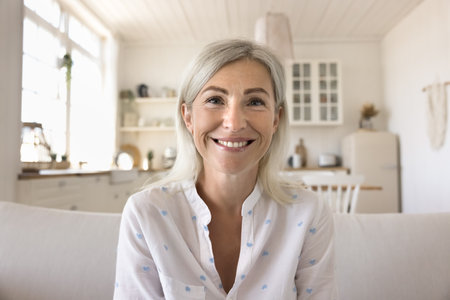 Positive pretty blonde mature woman home head shot portrait. Happy senior lady talking on online video conference call, looking at camera with toothy smile, sitting on couch with kitchen in backgroundの写真素材