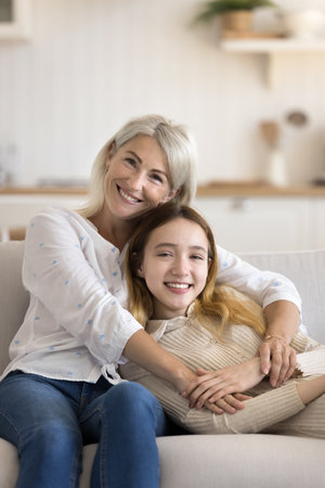 Happy mature grandma and adorable teenage grandkid girl resting on home couch, hugging with love,...