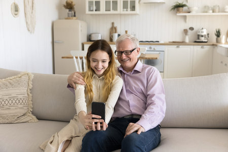 Cheerful grandpa and positive teenage kid girl taking family portrait on mobile phone, using...