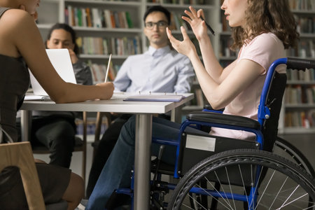 Team of college students and speaker with disability meeting in library, discussing group homework task, class project. Young college girl using wheelchair, talking to classmates. Cropped shotの写真素材