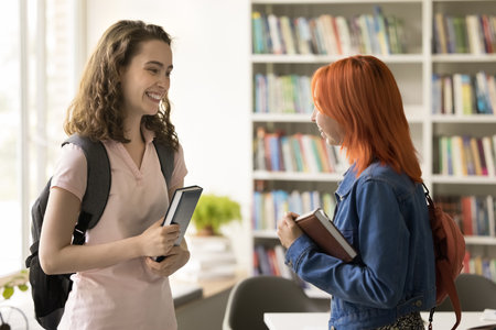 Two happy college mate girls meeting in campus library, standing together, talking, laughing,...