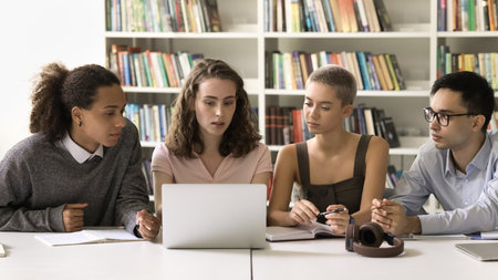 Diverse team of university students watching online learning content, webinar, lecture on Internet on laptop, sitting at computer in college library, working together on homework, research studyの写真素材
