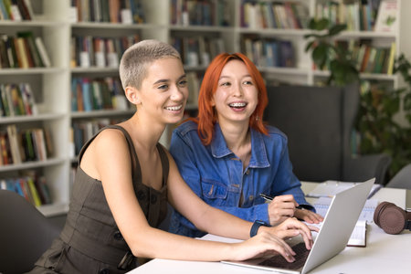 Cheerful college girl friends with teeth brackets working on class project in library together, studying in campus, sitting at table, typing on laptop, smiling, laughing, looking awayの写真素材