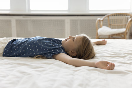 Positive peaceful relaxed little girl kid resting on bed with white linen, lying on comfortable mattress with open arms and closed eyes, sleeping in bedroom at daytimeの写真素材
