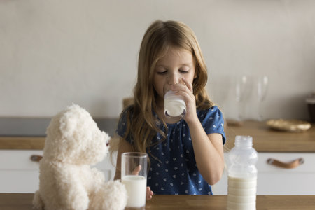 Pretty adorable preschool girl drinking milk in home kitchen, getting calcium, probiotics, holding glass, standing at table with bottle, teddy bear toy, enjoying healthy diet, natural nutritionの写真素材