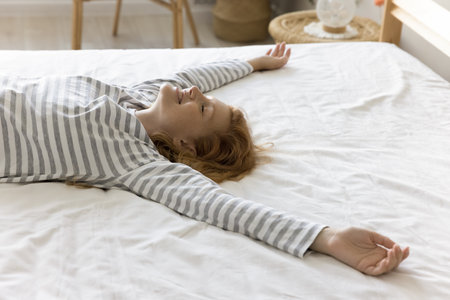 Positive relaxed pretty young woman resting on bed with open arms and closed eyes, lying on back on...