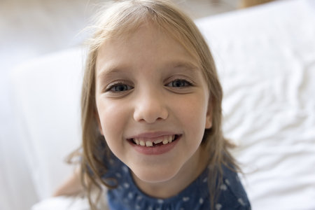Positive charming pretty preschool girl with fair hair, blue eyes, tooth gap looking at camera with...