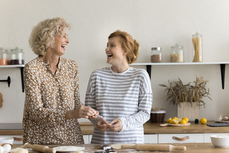 Joyful excited mom and adult daughter baking sweet homemade pastry snacks, cookies, biscuits, shaping, rolling dough, talking, discussing family recipe, smiling, laughing, having funの写真素材
