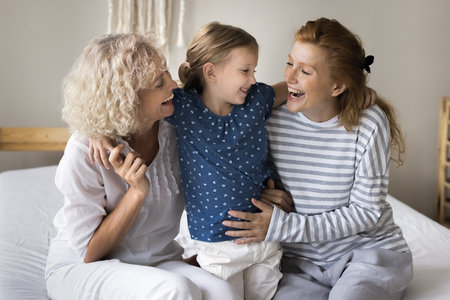 Cheerful little child, mom and grandma having fun on home bed, hugging with love, tenderness,...