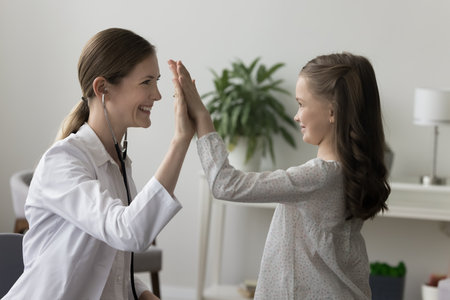 Cheerful pediatrician woman giving high five to little patient girl after using stethoscope, checking heartbeat, breath, smiling, laughing, thanking kid for trust, giving supportの写真素材