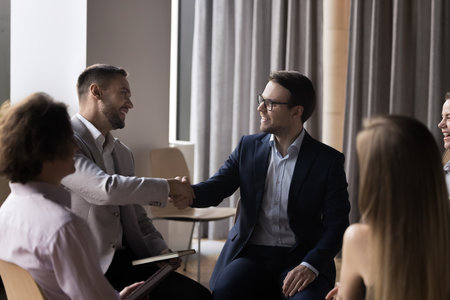 Two businessmen in formal suits shaking hands, start group formal meeting in office, gathered to discuss business, making profitable commercial deal. Express respect, negotiation process, etiquetteの写真素材