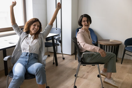 Two excited joyful business colleagues celebrating teamwork success, riding, wheeling office chairs,...