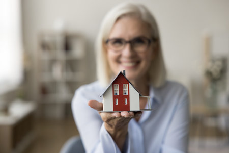 Tiny house model on hand of real estate agent woman looking at camera, smiling in blurred background. Senior realtor offering property buying, rent of apartment, mortgage consultationの写真素材