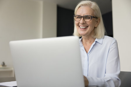 Happy pretty mature older business woman in elegant eye glasses working at laptop, typing, talking on online video call, laughing, smiling, using computer for job communicationの写真素材