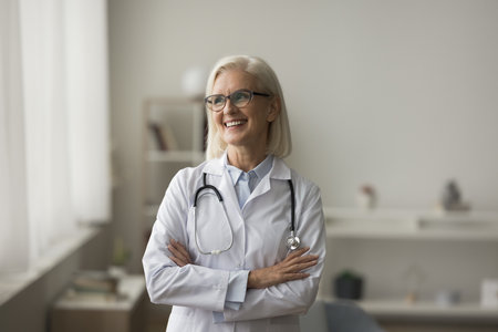 Positive older mature practitioner woman in glasses posing indoors, looking at window away, thinking, smiling. Senior medical professional, doctor in white uniform casual portraitの写真素材