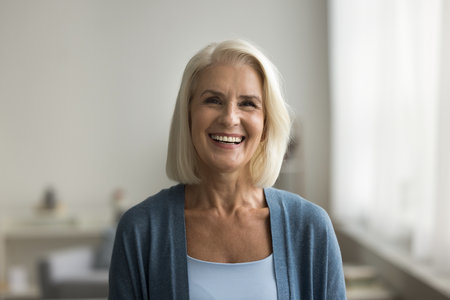 Happy blonde senior retired woman looking at camera with perfect toothy smile, posing at home,...
