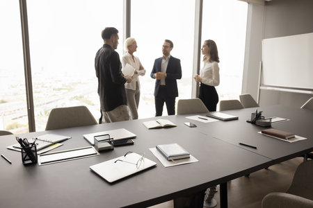 Business team of four discussing work project in office conference room, talking, standing in background at large window, white board, meeting table with stationery, notebooks, empty chairsの写真素材
