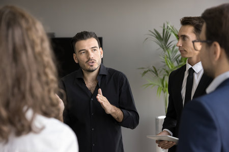 Handsome business leader man talking to group of partners on meeting, telling ideas, presenting project. Team of professionals standing indoors, listening to speakerの写真素材