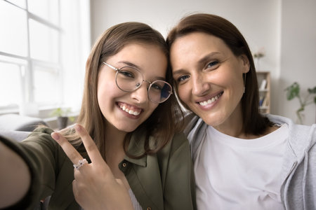 Happy loving mom and cheerful pretty teenage kid girl taking family selfie together, posing for self portrait at home, holding gadget with camera, smartphone in handの写真素材
