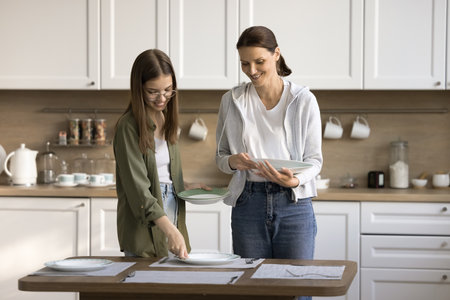 Happy mother and teen daughter child decorating table for dinner, placing dich, plates, talking, chatting, smiling, laughing, enjoying household activities. Kid helping mom with domestic choresの写真素材