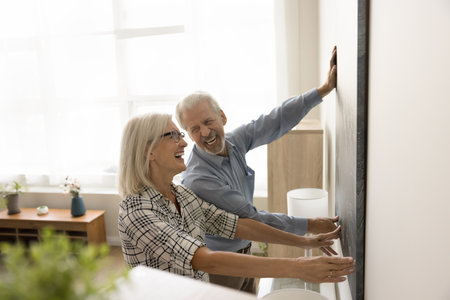 Joyful positive retired husband and wife enjoying interior decoration work, hanging large artwork on wall, holding picture together, smiling, laughing, having in new apartmentの写真素材