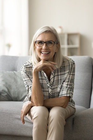 Cheerful beautiful senior lady in trendy glasses leaning chin on hand, sitting on home sofa, looking at camera with toothy smile, talking on online video conference call. Vertical shotの写真素材