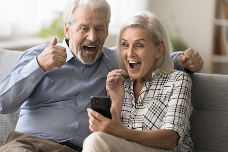 Excited surprised retired couple holding mobile phone, staring at gadget screen, shouting for joy with winner hands, celebrating victory, success, win, enjoying leisure, entertainment at homeの写真素材