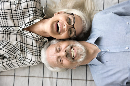 Cheerful relaxed senior couple faces top view above shot. Happy carefree retired husband and wife in love lying on backs with head touches, looking at camera, smiling, laughing. Close portraitの写真素材