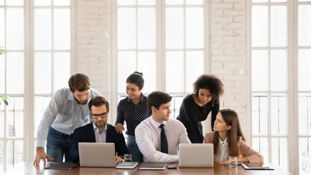 Smiling diverse employees sit at desk work in groups on computers brainstorming in office together, happy motivated young multiracial businesspeople cooperate using laptops, teamwork conceptの写真素材