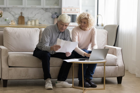 Serious senior retired couple counting domestic budget, reading paper documents on couch in living...