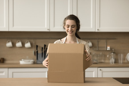 Happy beautiful young customer woman opening package, unpacking cardboard box, parcel on kitchen...