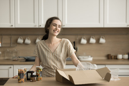 Happy pretty adult girl unpacking cardboard box with canned vegetables in kitchen, getting grocery purchase from supermarket, standing at table with glass jars, smiling, laughingの写真素材
