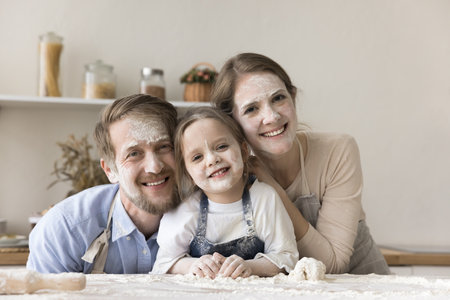 Happy young mom, dad and toddler kid in aprons with funny floury facial masks cooking dessert in home kitchen, having fun, posing at table with heap of flour, looking at camera, laughingの写真素材