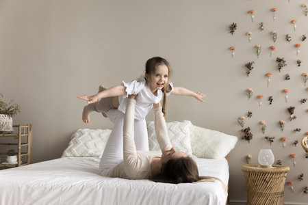 Sportive strong mom lifting kid in air, supporting child with arms and ankles, lying on bed. Happy excited girl playing airplane held by mom, flying with open hands, exercising, doing acroyogaの写真素材