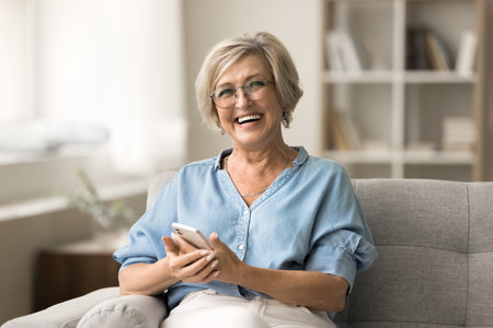 Cheerful pretty older retired woman in stylish glasses holding smartphone on couch, looking at...
