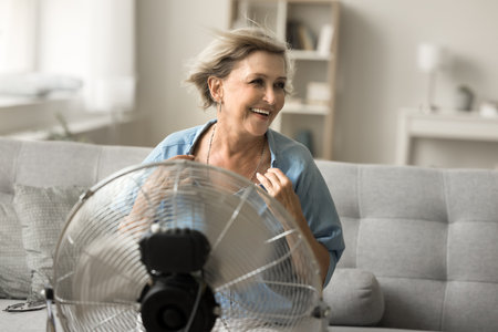 Cheerful carefree senior woman enjoying freshness from electric blower, ventilator, cooler, sitting at fresh air blowing from fan, using home appliance, equipment for coolingの写真素材