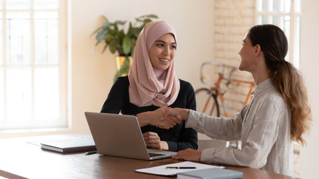 Young middle eastern ethnicity and mixed-race businesswomen colleagues sit at desk in office shaking hands express trust, appreciations for work done after teamwork, executive welcoming client conceptの写真素材