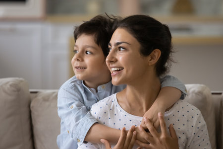Head shot smiling dreamy Indian mother piggy backing 5s son, looking to aside in distance, visualizing good future, planning, adorable boy child hugging loving young mom, motherhood conceptの写真素材