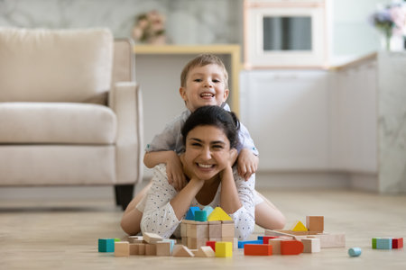 Portrait happy Indian mother with Caucasian son having fun, playing with toys on warm floor at home, smiling 7s boy sitting on young mom back, looking at camera, family enjoying leisure timeの写真素材
