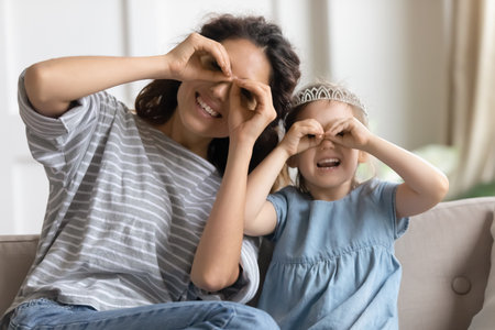 Portrait of smiling young mother and cute little preschooler daughter sit on couch make funny faces together, happy millennial mom or nanny posing with small girl child enjoy weekend at home togetherの写真素材