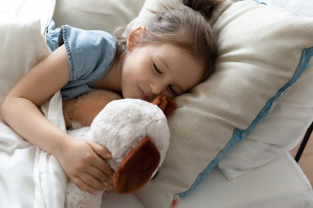Cute little preschooler girl lying in cozy bed hugging taking nap with favorite plush toy, small kid sleep relax in comfortable bedroom cuddling with fluffy teddy bear at home, relaxation conceptの写真素材
