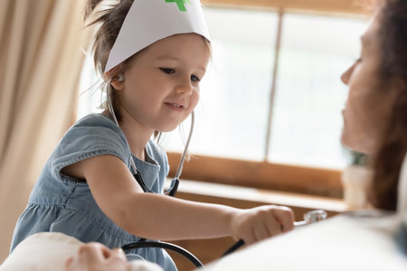 Little preschooler girl wearing medical uniform appliances playing nurse patient game with mom or nanny, small cute child acting as doctor listen to mother chest, engaged in funny activity at homeの写真素材