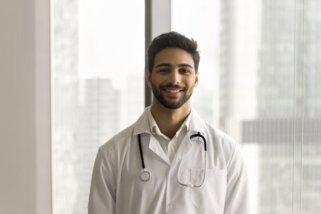 Happy handsome young Arab doctor man wearing white uniform coat and stethoscope posing for shooting, looking at camera, standing at large window in clinic office. Healthcare professional portraitの写真素材