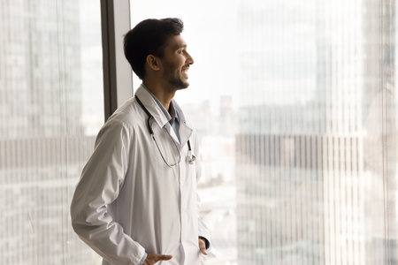 Happy successful Indian doctor man looking at city out of window, standing in hospital office indoors, smiling, laughing, feeling satisfied about medical job, profession, thinking on careerの写真素材