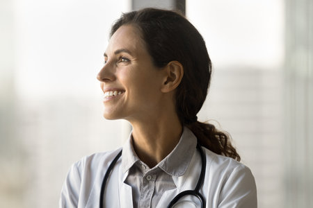 Happy pretty young doctor woman with stethoscope on her neck looking away with toothy smile. Positive therapist standing at window glass, thinking on successful medical careerの写真素材