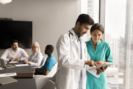 Two focused diverse doctor colleagues using tablet on corporate meeting, discussing healthcare...