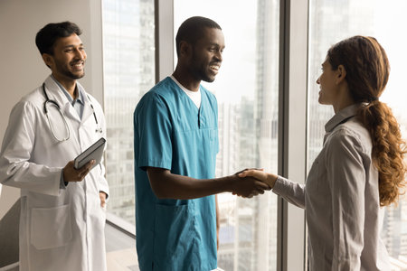Positive female doctor shaking hands with young African American surgeon in scrubs, thanking colleague for good successful job, cooperation, expressing professional respectの写真素材