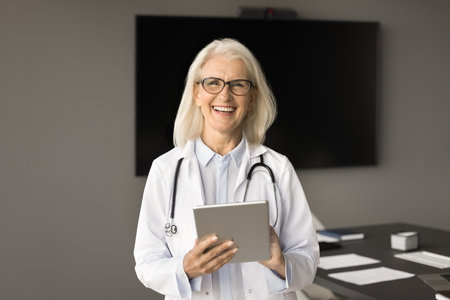Happy confident doctor woman in glasses using medical service on digital tablet, enjoying job online communication, holding gadget, looking at camera, smiling, laughing with boardroom in backgroundの写真素材